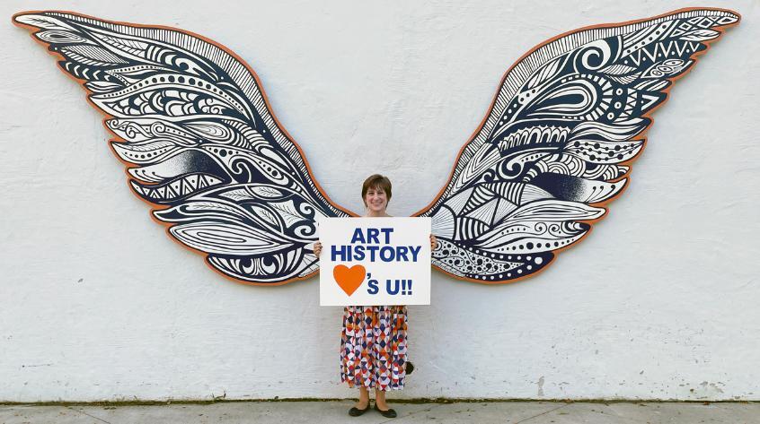 a person standing in front of wings holding a sign that says Art History loves you!
